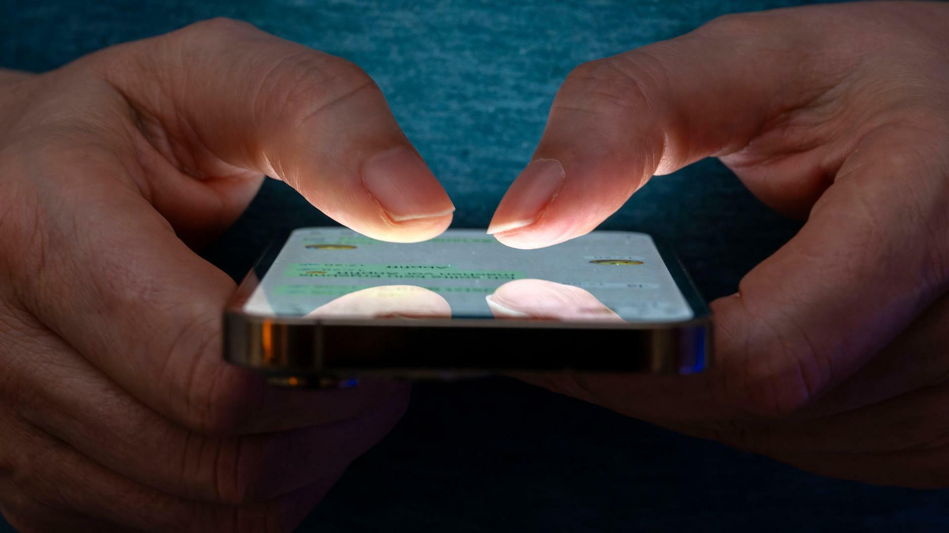 Eine Frau tippt auf einem Smartphone-Screen. Das Bild zeigt eine Person, die in einer schlecht beleuchteten Umgebung ein Smartphone benutzt. Die Beleuchtung hebt die Finger des Benutzers und den Bildschirm des Smartphones hervor.