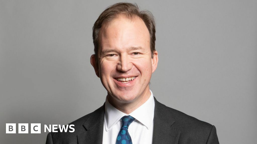 A portrait photograph of MP Jesse Norman. He is wearing a grey suit jacket and white shirt with a blue spotty tie. He is stood against a grey backdrop.