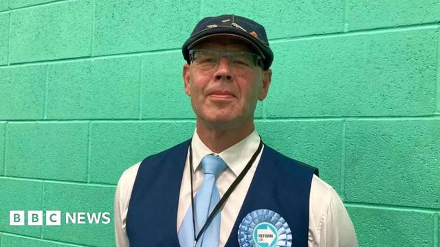 A man in a white shirt with blue waistcoat and blue cap and light blue tie and light blue Reform UK rosette standing against a green wall