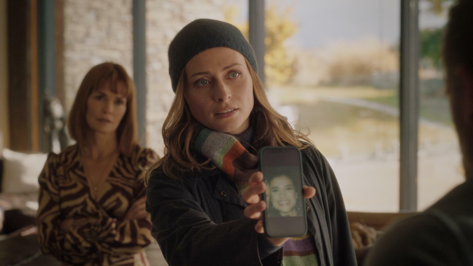 "Queenstown Murders – Zum Sterben schön - Am Abgrund": Detective Sergeant Anais Mallory (Chelsie Preston Crayford) zeigt Sebastian van Heusen (Mike Edward) ein Handy-Display mit dem Foto einer Frau. Er ist nur ansatzweise im Profil zu sehen. Aus dem Hintergrund beobachtet Marijke van Heusen (Nathalie Boltt) den Vorgang. Sie wirkt verärgert.