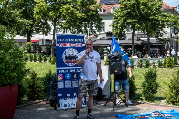 Bild einer rassistischen Kundgebung am 29.06.24 in Bad Oeynhausen. In der Bildmitte steht Daniel Kokott im Shirt aus dem Shop der Identitären Bewegung und kurzer Hose im Tarnmuster. Er spricht in ein Mikrofon. Im Hintergrund ist ein Aufsteller der rechten Gruppe "Bad Oeynhausen erhebt sich" zu sehen.