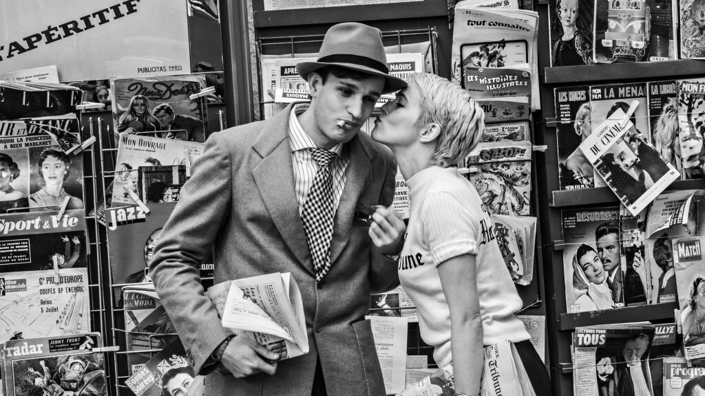 Zoey Deutch kisses Aubry Dullin on the cheek in front of a newsstand in a black-and-white still from 'Nouvelle Vague'.