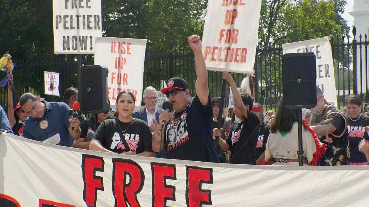 A group of demonstrators rallying behind a large banner reading “Free Leonard Peltier,” holding signs that say “Free Peltier Now!” and “Rise Up for Peltier.” 