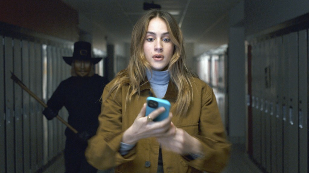 Nell Verlaque looks at her phone with shock in a dark school hallway someone in a pilgrim mask follows her with an ax in 'Thanksgiving' (2023).