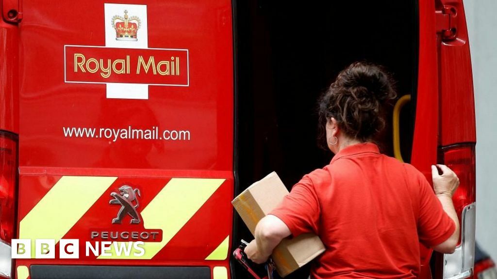 A female postal worker holds a parcel in one arm and closes a door on the back of a red Royal Mail van with the other