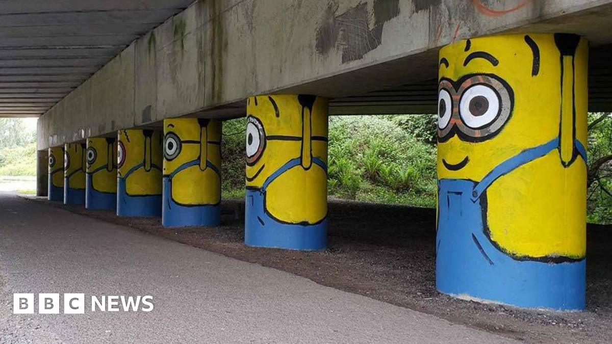 A row of eight painted stanchions under a motorway bridge, looking like yellow minions wearing blue dungarees and holding up the bridge