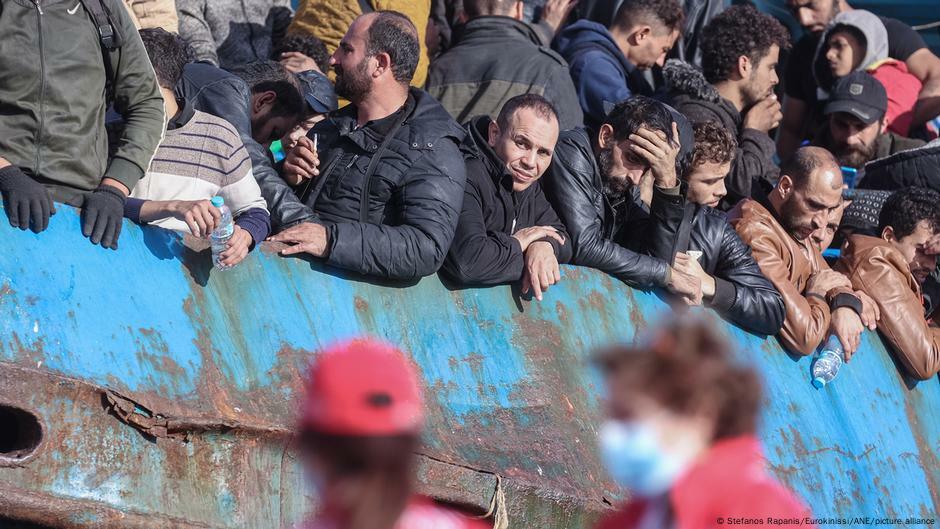 Many men in winter jackets crowded close together, looking down over the blue-painted side of a ship. The photo was taken in Chania, Crete, in November 2022.