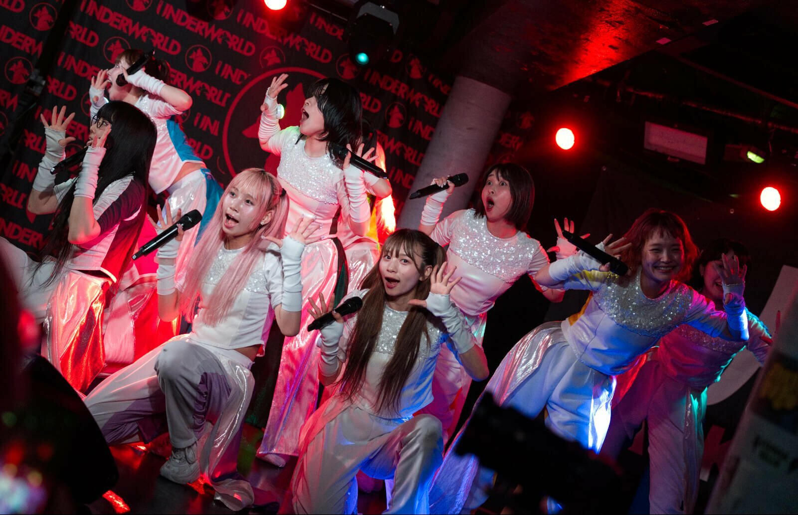 The photo depicts a moment where the idol group GANG PARADE, consisting of 11 members, are at the end of a song and posing. All members are dressed in white outfits with on the back a bright colour, it is a different colour for everyone.