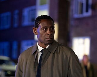 A person filming Pierre in a brown trench coat and tie stands in a nighttime urban scene with blurred buildings and illuminated windows in the background, alongside a white car and another figure.