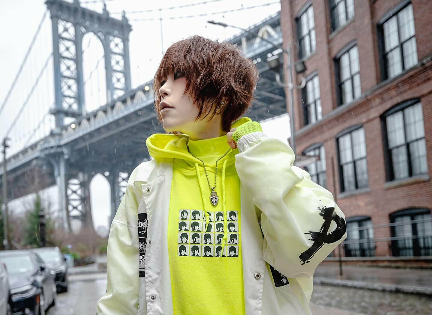 Photo of NANO standing in a street in New York on a rainy day. Behind NANO is a cable bridge and she is dressed in a white jacket over a neon yellow hoodie. Photo by Joey (@evillittlejawa)