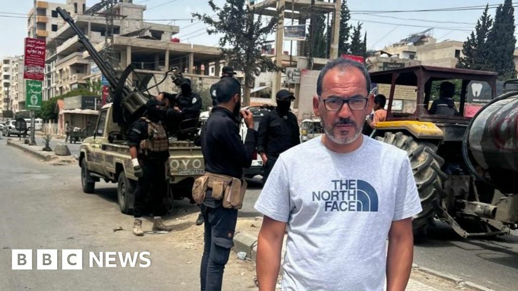 Feras Kilani stands on a street in Damascus, Syria. In the background, Syrian security forces are deployed with a truck-mounted anti-aircraft gun (7 May 2025)