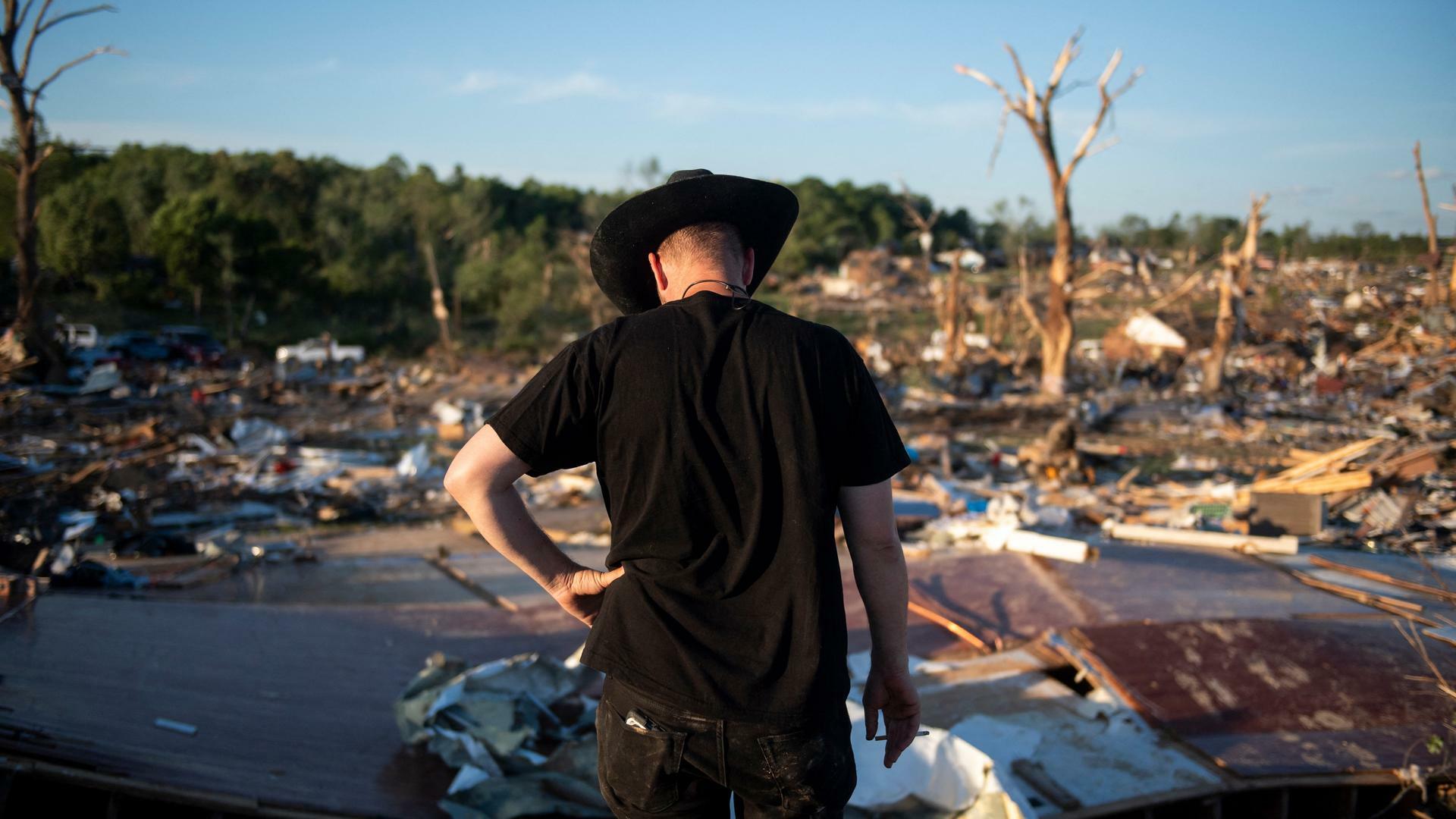 Ein Mann mit schwarzem Cowboyhut steht mit gesenktem Kopf vor den Überresten eines durch Tornados zerstörten Gebäudes.
