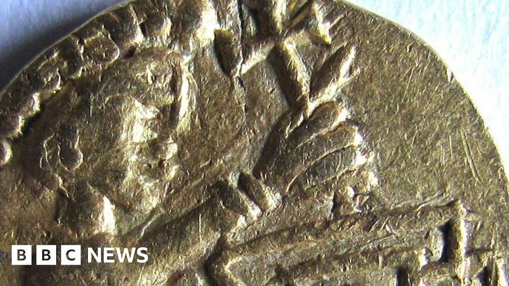 A close up of a 7th Century gold coin. It shows a male figure with an oversized head, wearing a Roman-style headdress or diadem. Its left arm is raised up and appears to be holding a cross.
