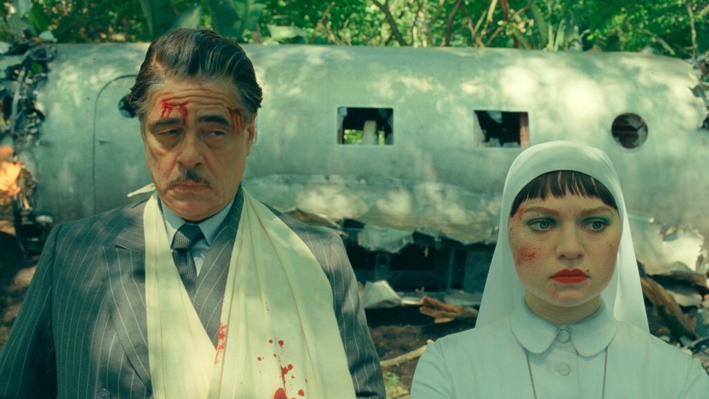 Benicio Del Toro and Mia Threapleton stand in front of a crashed airplane in 'The Phoenician Scheme' (2025).
