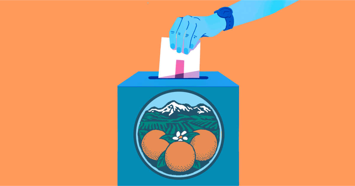 A person's hand drops a ballot into a ballot box with oranges and a view to snow-capped mountains