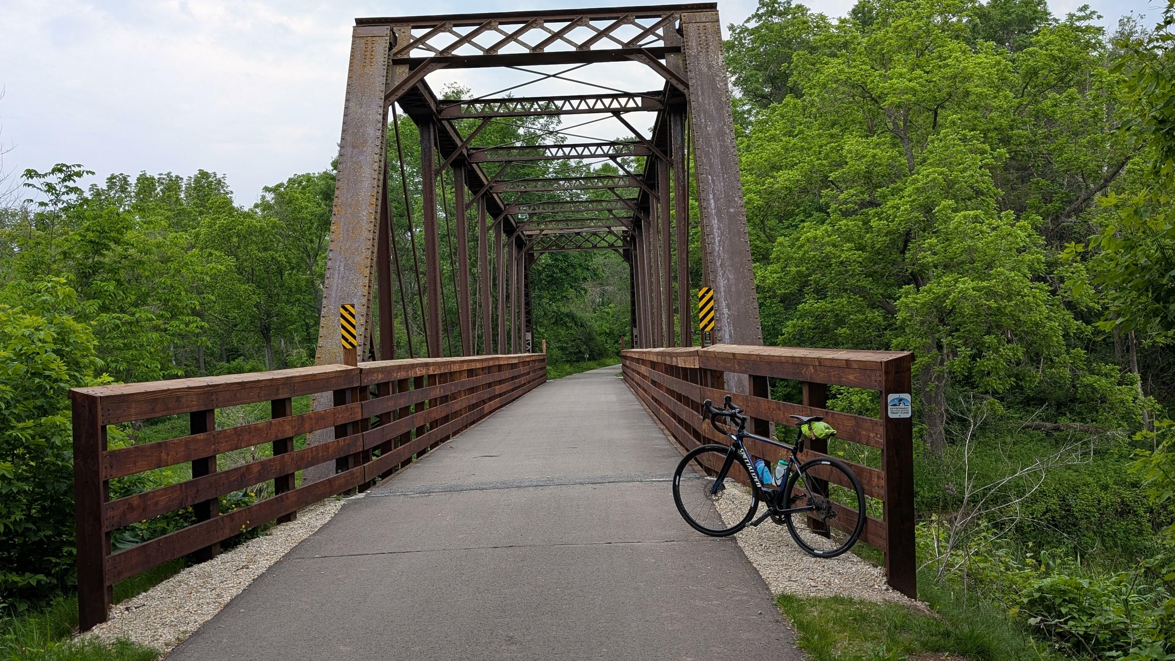 Blue and black Roubaix bicycle leaning on the wooden railing of a converted rusty colored steel truss railroad bridge with an asphalt bike trail deck over the Root River, surrounded by trees on a partly cloudy day.