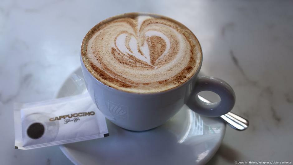 A cup of cappucino on a table, with spoon and sachet of sugar draped on the saucer