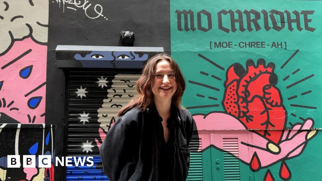 Katie Forbes stands in front of a colourful street mural of a heart being held in a hand. She has long brown hair and a black jacket.