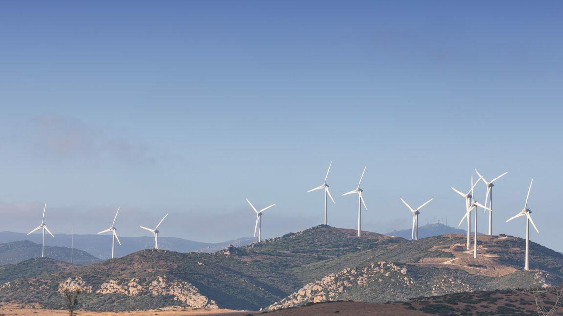 Image of a dry, brown hillside with lots of small scrub, and white wind turbines in the background.