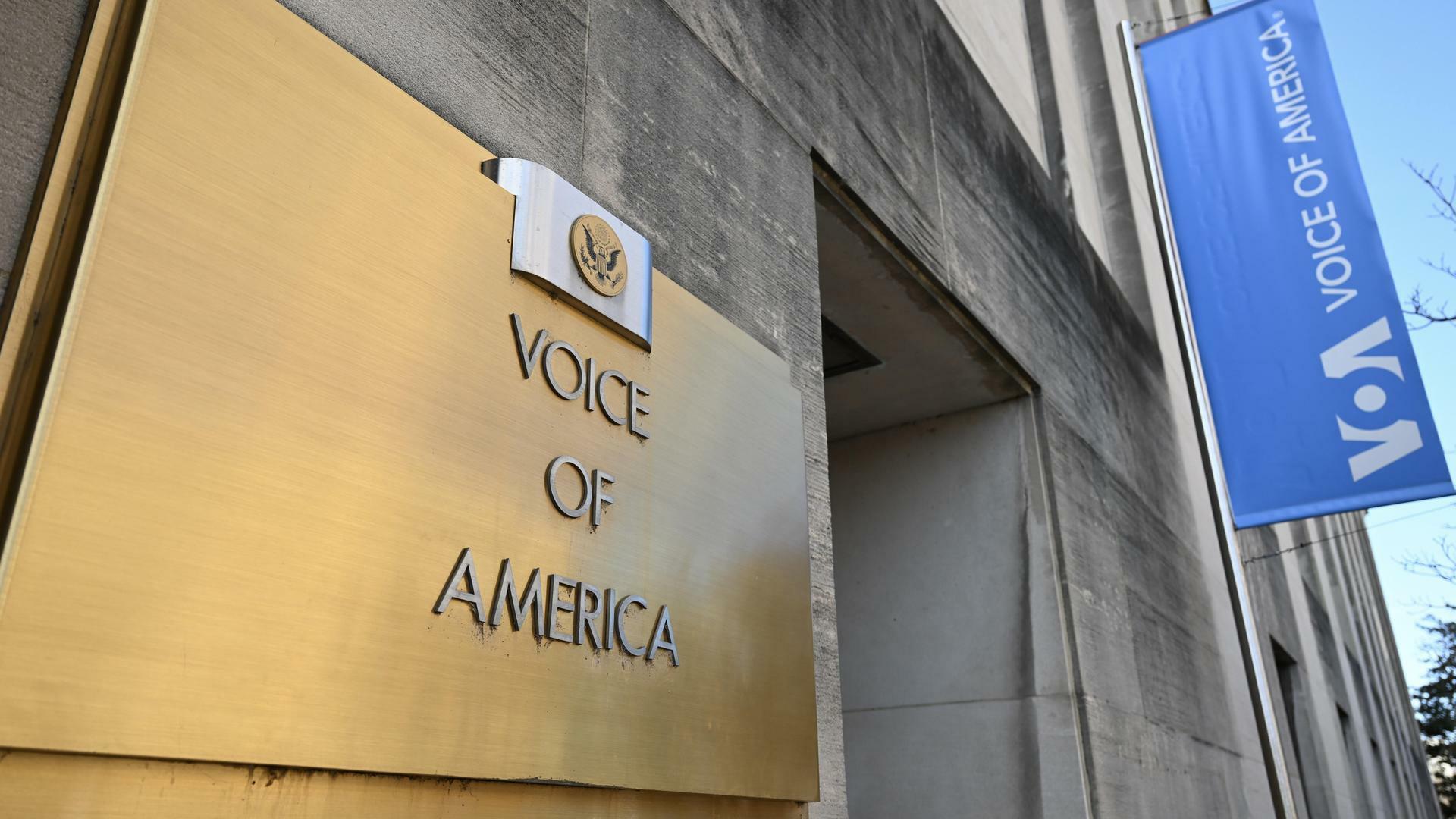 Blick von schräg unten auf das Schild am Eingang eines Gebäudes von Voice of America, im Hintergrund weht eine bleue Fahne mit dem Logo des Senders.
