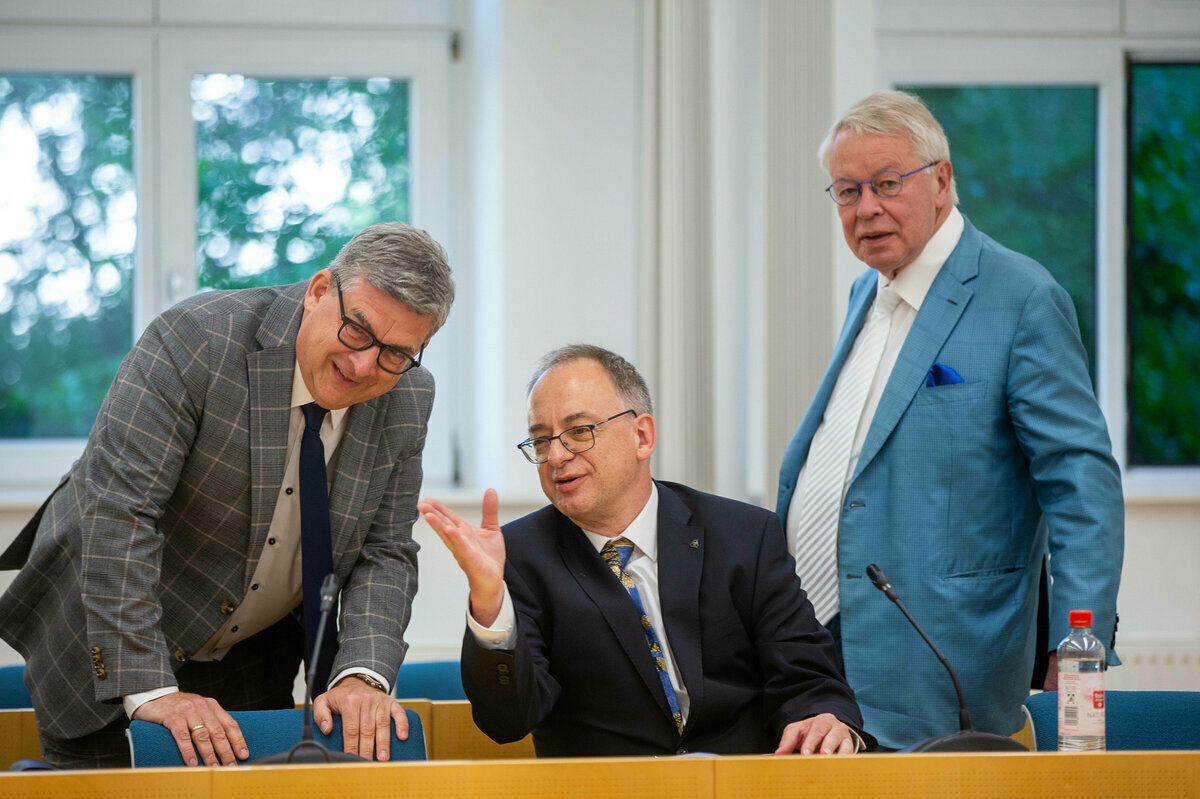 Drei Männer im Gerichtssaal, einer sitzt, die beiden anderen Männer stehen neben ihm und hören ihm zu