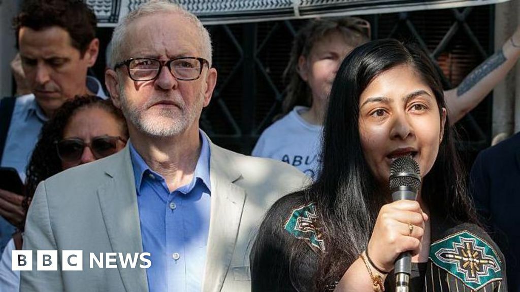 Jeremy Corbyn and Zarah Sultana stand among a group of people at a protest. Corbyn wears a light-colored blazer over a blue shirt. Sultana, dressed in a black outfit with colorful embroidery, holds a microphone.