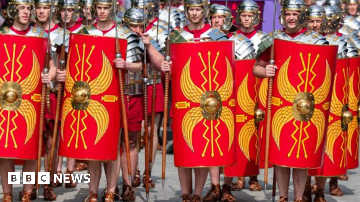 A large group of men dressed as ancient Roman legionaries. They are dressed in silver-coloured armour, including helmets, over red tunics. They are carrying spears and red shields with a yellow design on them. On their feet are leather sandals, tied up around their ankles. 