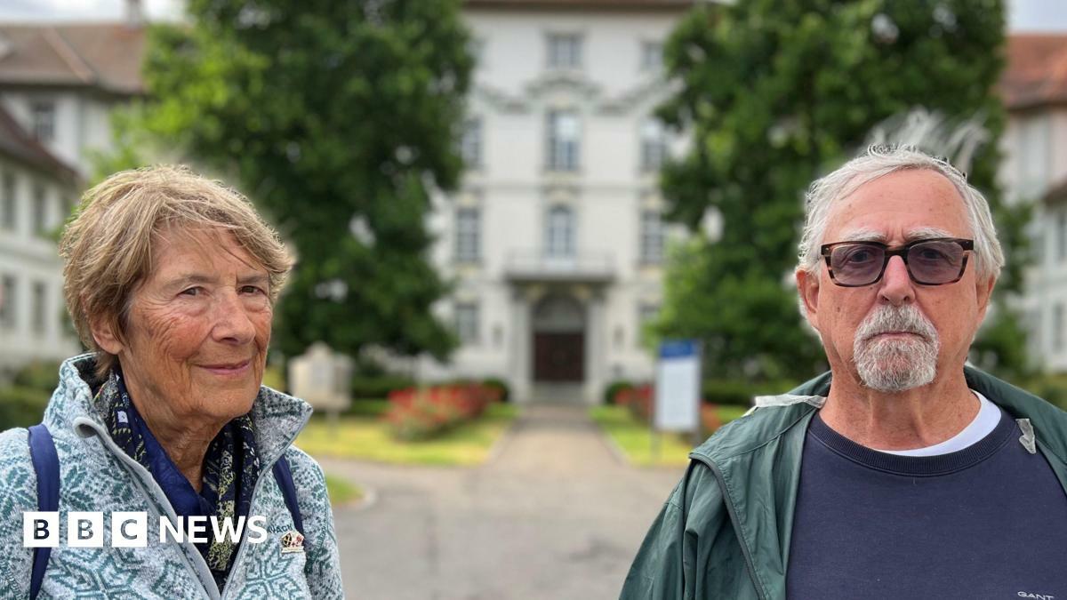 Lola and David are standing outdoors in front of a large, white building with a red roof and multiple windows.
