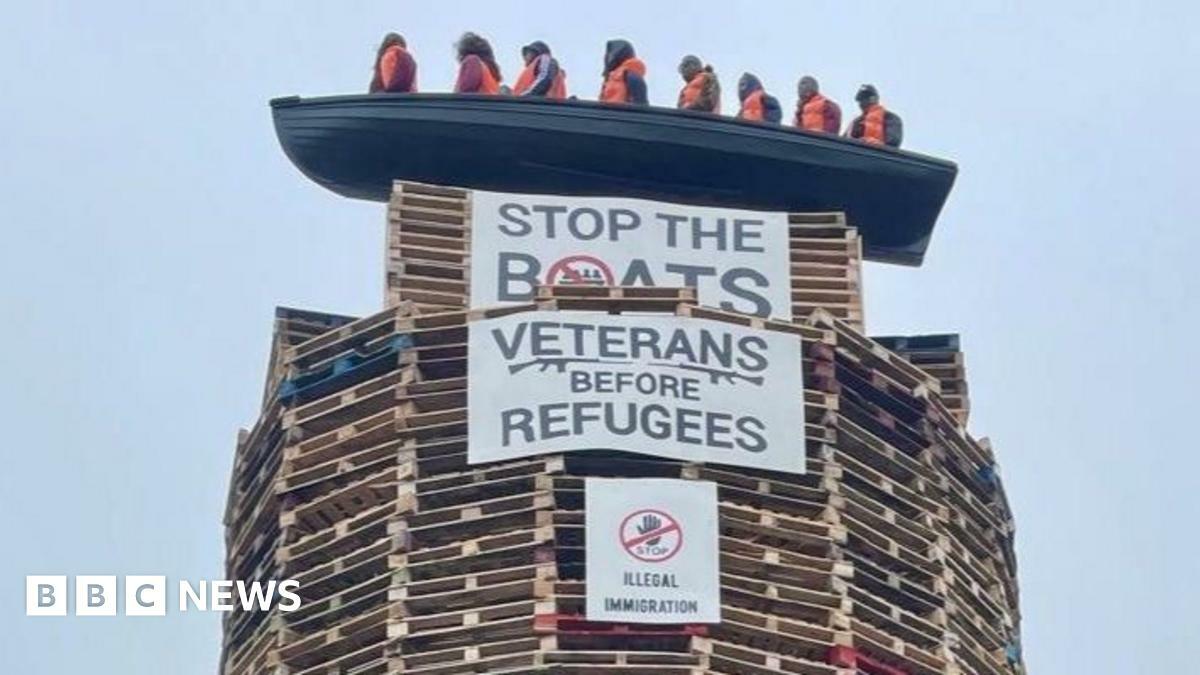 The picture shows a model of a small boat with figures inside on top of a tall bonfire constructed mainly from pallets. Signs saying "Stop The Boats" and "Veterans Before Refugees" are on the bonfire.