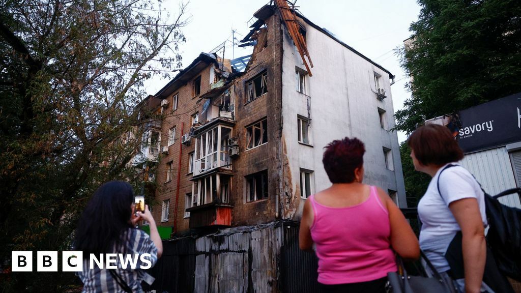 People stand outside looking at a residential building in Kyiv damaged in a Russian drone attack, with one taking a photo on their phone. The building is visibly damaged with a large hole on the top floor and trees line the street, in Kyiv on 10 July. People stand outside looking at a residential building in Kyiv damaged in a Russian drone attack, with one taking a photo on their phone. The building is visibly damaged with a large hole on the top floor and trees line the street, in Kyiv on 10 July.