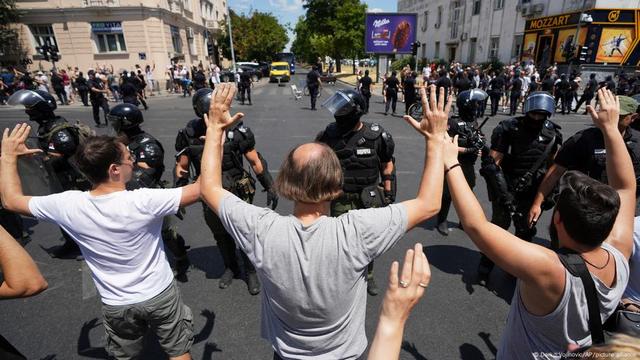 Civilians hold up their hands as they face a line of police officers in riot gear at an intersection. Other civilians and police officers face off in a similar way on the other side of the intersection in the background. Belgrade, Serbia, Monday, 30 June 2025. | Image: Darko Vojinovic/AP/picture alliance