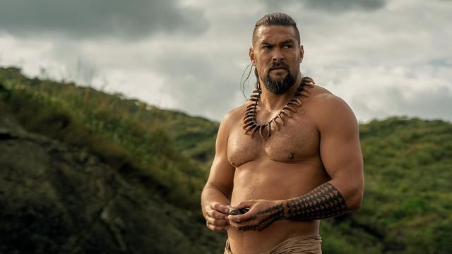 Jason Momoa, torse nu et portant un collier de petits os, incarne un chef de guerre hawaïen devant une forêt verdoyante.