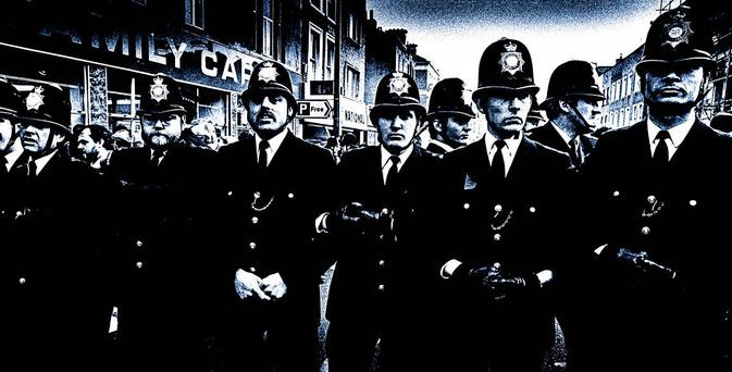 High-contrast, black-and-white photograph of a row of British police officers forming a barrier.