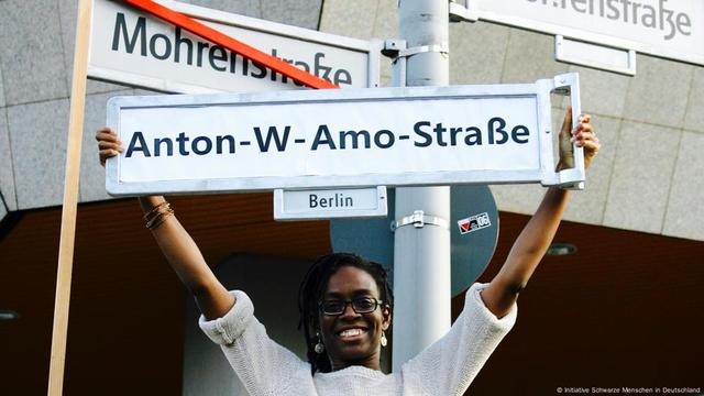 Eine Frau hält lächelnd das Straßenschild mit der Aufschrift "Anton-W-Amo-Straße" hoch.