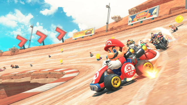 Mario races in a red vehicle while making a turn on a desert race track with several other racers behind him.