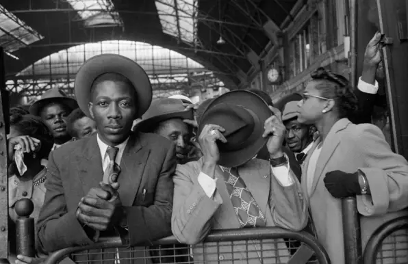 A black and white photograph showing several people of West Indian descent, dressed smartly in suits, ties and wearing hats in a 1950s fashion, against railings in Victoria Station.