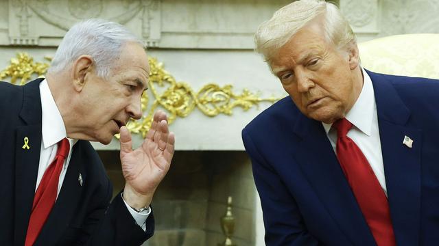 Two older men in suits with red ties sit close, one whispering to the other against a decorative white and gold background, suggesting a serious conversation in a formal setting.