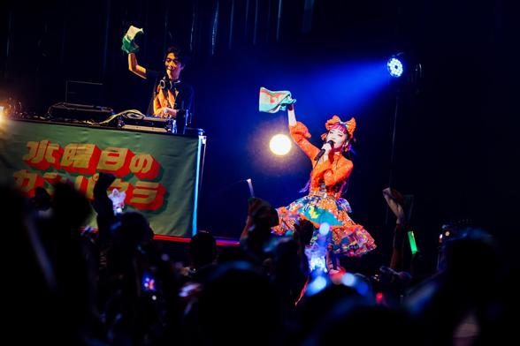 Photo of Wednesday Campanella at Asakusa Public Hall in Tokyo in April 2025. The photo captures a lively scene of a performance on a brightly lit stage. On the stage, Utaha is dressed in a colourful outfit and Kenmochi is on hte background on his DJ booth. Both are waving with a towel/muffler. Photo by Masato Yokoyama, courtesy of Tsubasa Records Photo of Wednesday Campanella at Asakusa Public Hall in Tokyo in April 2025. The photo captures a lively scene of a performance on a brightly lit stage. On the stage, Utaha is dressed in a colourful outfit and Kenmochi is on hte background on his DJ booth. Both are waving with a towel/muffler. Photo by Masato Yokoyama, courtesy of Tsubasa Records