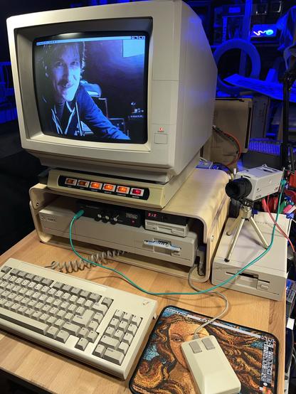 Photo of an Amiga 1000 with keyboard and mouse, and a monitor on top and a small black box in between with three knobs. A small video camera sits atop an external floppy disk on the right. In the screen is a picture of the author as digitized with the Frame Grabber. 
