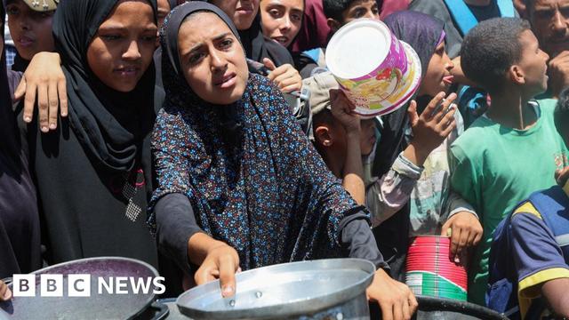 Two women, who are wearing black and have pained looks on their faces, stand at the front of a crowd and hold out pans at a charity kitchen in Nuseirat, Gaza