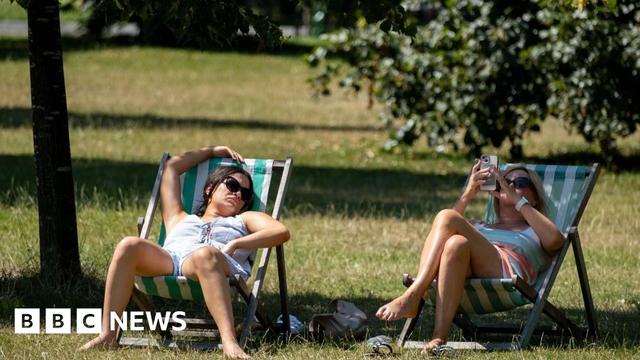 Two women sitting under tree shade on a noticeably hot day in a park in green and white striped deckchairs. The woman on the left has dark hair and sunglasses and appears to be asleep, with her right arm rest above her head on top of the deckchair. The woman on the right is blonde and is also wearing sunglasses and is looking at her phone.