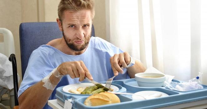 Ein Patient in einem Krankenhaus macht sich mit Messer und Kabel an dem Essen auf einem Tablett zu schaffen. Er rümpft dabei die Nase.