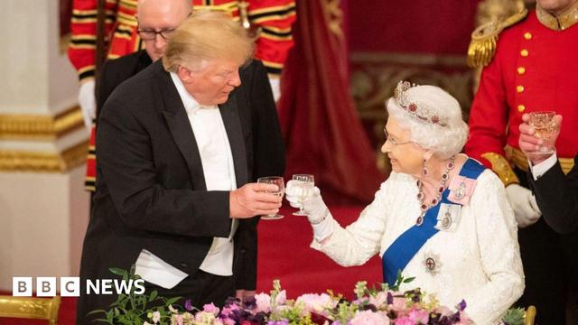 Donald Trump raises a glass with Queen Elizabeth II during a state banquet as part of his 2019 visit to the UK. He wears a formal black suit, white shirt and bow tie, while she wears a white, long-sleeved dress, silver crown and a ruby and diamond necklace. The pair are touching wine glasses and appear to be attended by two footmen.