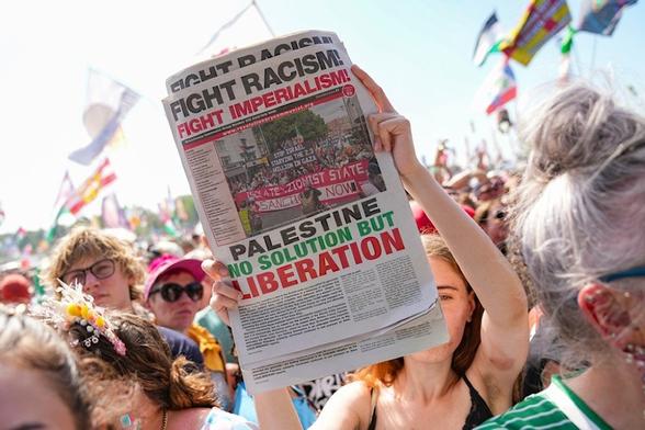 A festivalgoer holds a newspaper that reads "Fight Racism! Fight Imperialism! Palestine No Solution But Liberation" during the performance of hip hop trio Kneecap at Glastonbury Festival in Worthy Farm, Somerset, England, Saturday, June 28, 2025 (AP)