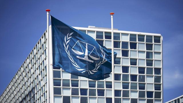 Außenansicht des Internationalen Strafgerichtshofs (IStGH). Davor weht die Flagge des IStGH, eine Waage mit zwei Waagschalen, die von zwei Olivenzweigen umschlossen wird.