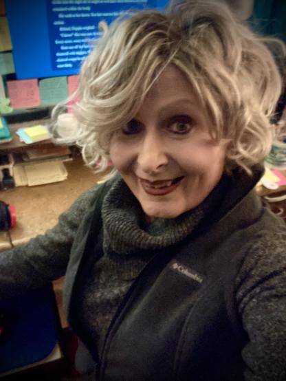 A blonde woman is facing to the left, but looking at the camera. She's wearing a gray sweater, a vest over that. Behind her is a desk, the computer on and various colored sticky notes around its edge. She appears happy and satisfied.
