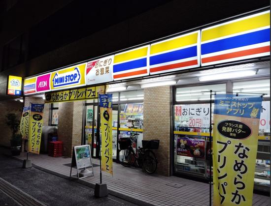 A nighttime photo of the author's neighborhood Mini-Stop. The long neon sign of three colored lines, (from top to bottom, yellow, blue and red, the yellow and blue thicker than the red), is at the top, disrupted by a square of a pink AEON sign, the larger company that owns Mini-Stop. The Mini-Stop logo is a line illustration representing a store with the words 'Mini-Stop' in blue. Many yellow vertical signs with vertical hiragana and katakana script are in from of the store. The wall parts of the store and the ground are tiny square cement tiles in a more brownish yellow color. The windows of the store show bright light inside and are themselves covered with many signs.