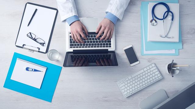 An overhead view of a doctor's desk featuring a laptop, sphygmomanometer, glasses, notepad, smartphone, and a stethoscope, illustrating a medical workspace. An overhead view of a doctor's desk featuring a laptop, sphygmomanometer, glasses, notepad, smartphone, and a stethoscope, illustrating a medical workspace.