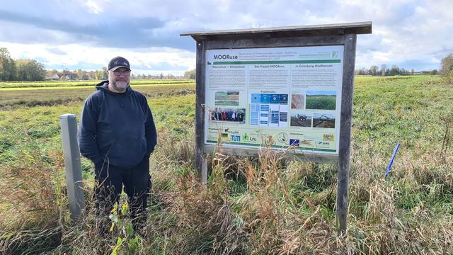 Mann steht auf einem Feld mit hohem Gras und einer Infotafel.