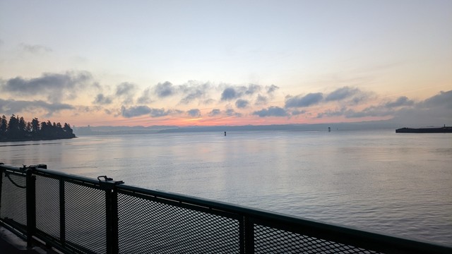 Sunrise, as seen from the MV Kaleetan, with its deck railing in frame, as it sets sail from Bainbridge Island to Seattle. There are wispy clouds near the horizon, which glows pink.